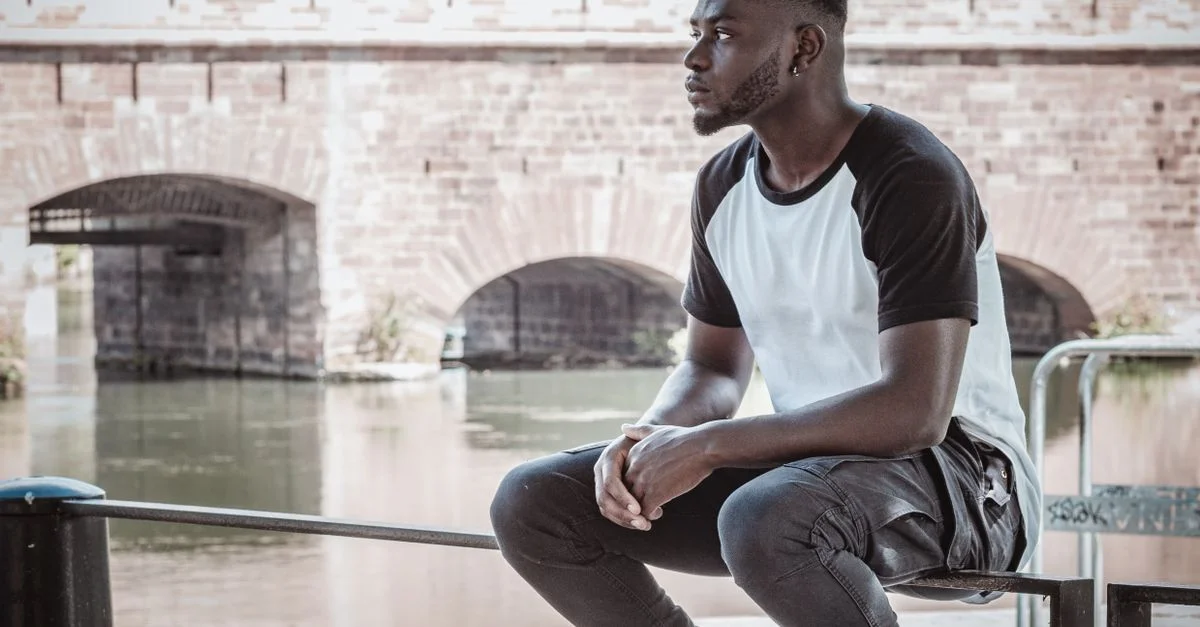 Jeune homme pensif assis près d'une rivière avec un vieux pont en pierre à Strasbourg, France.