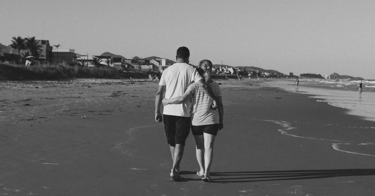 Photo en noir et blanc d'un couple marchant bras dessus bras dessous le long d'une plage tranquille.
