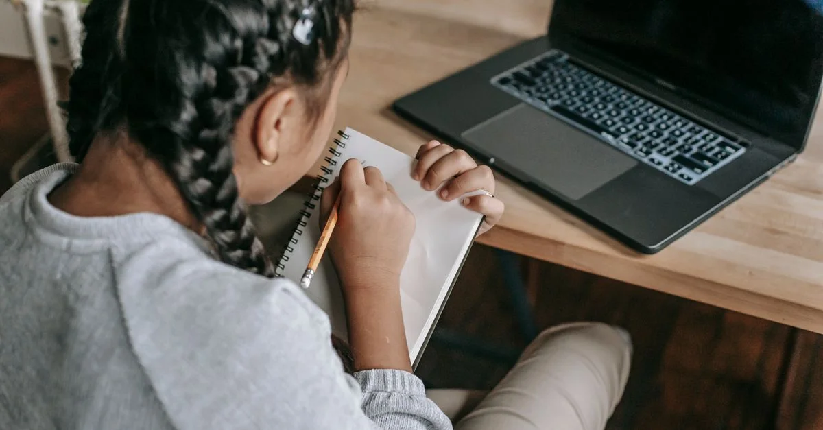 Jeune fille écrivant dans un carnet avec un ordinateur portable, montrant un apprentissage concentré.
