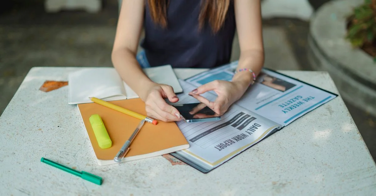 Jeune femme étudiant en plein air avec smartphone, carnet et surligneur, multitâchant entre études et communication.