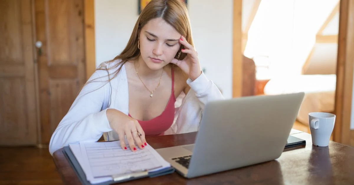 Jeune femme concentrée travaillant sur des documents et un ordinateur portable dans un cadre domestique confortable.