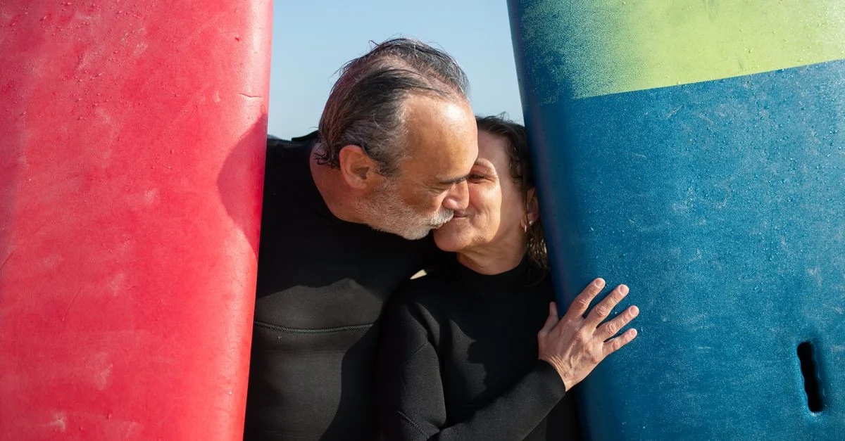 Couple senior en combinaisons de plongée s'embrassant entre des planches de surf, symbolisant l'amour au bord de la plage.
