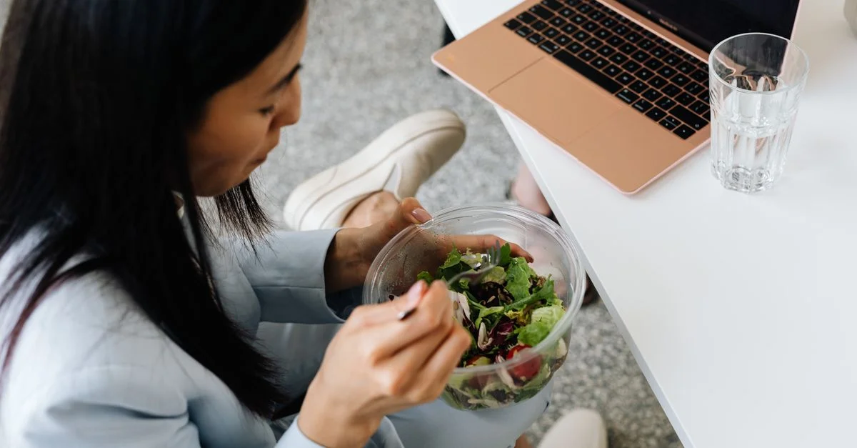 Femme mangeant une salade fraîche à son bureau avec un ordinateur portable à proximité, promouvant une alimentation saine au travail.