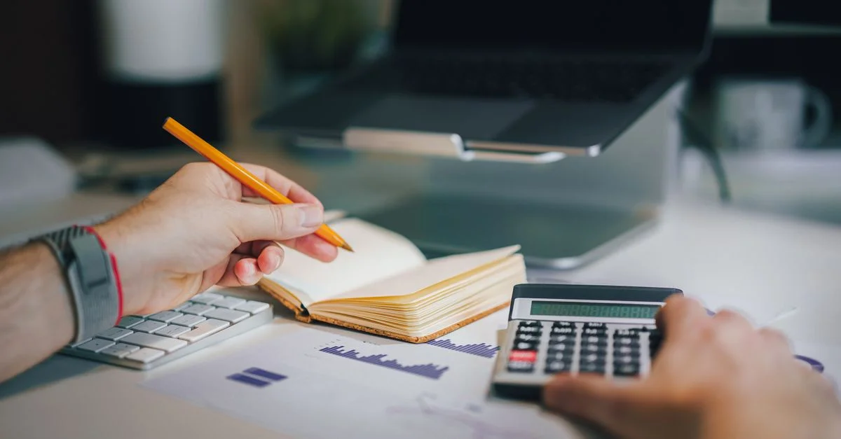 Mains utilisant une calculatrice et prenant des notes sur un bureau avec un ordinateur portable, mettant en avant l'analyse commerciale.