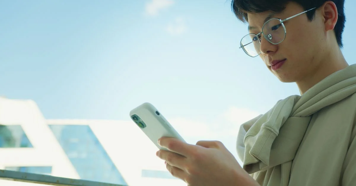 Un jeune adulte portant des lunettes utilise un smartphone en extérieur par une journée ensoleillée.