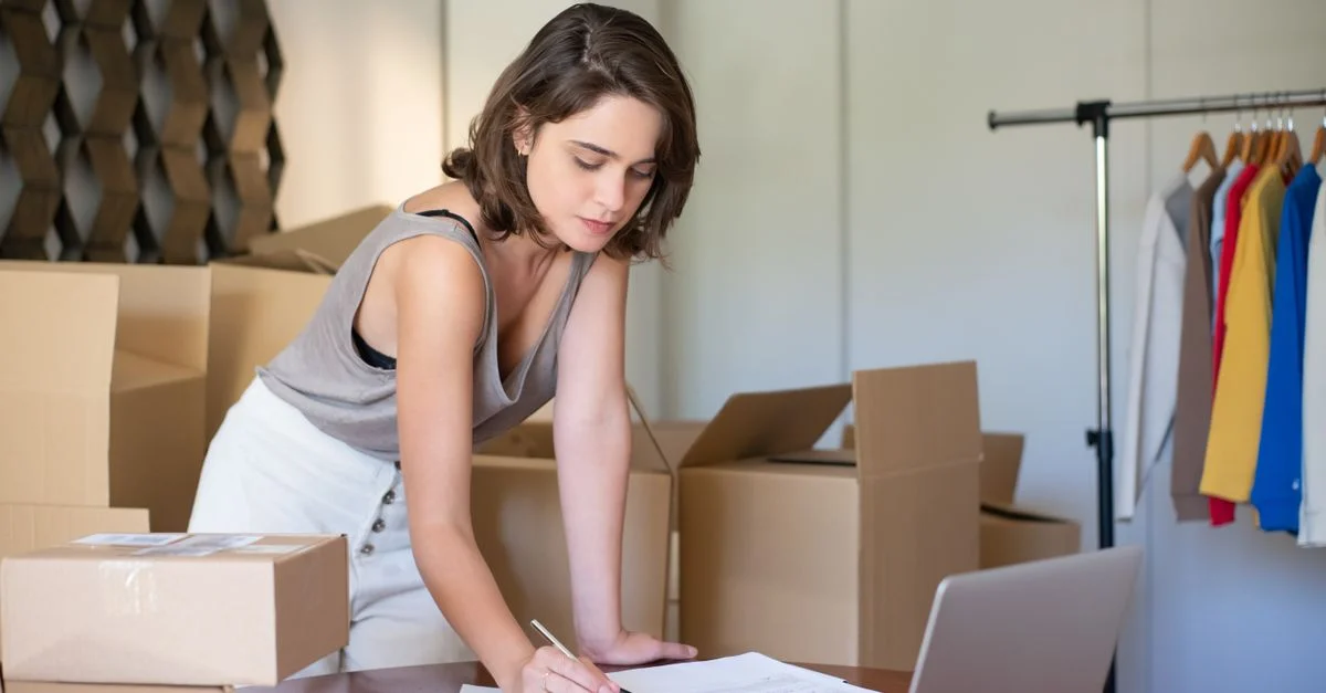Jeune femme gérant son entreprise de vêtements en ligne depuis son bureau à domicile avec des cartons et un ordinateur portable.