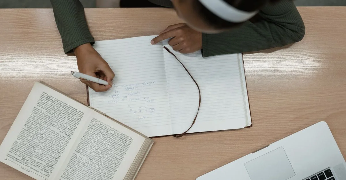 Vue de dessus d'une personne étudiant avec un livre ouvert et un ordinateur portable, prenant des notes dans un carnet.